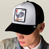 A person wearing a black and white trucker hat with a square patch featuring a rooster and the word 'cock'. The photo is cropped from the shoulders up against a plain light beige background.