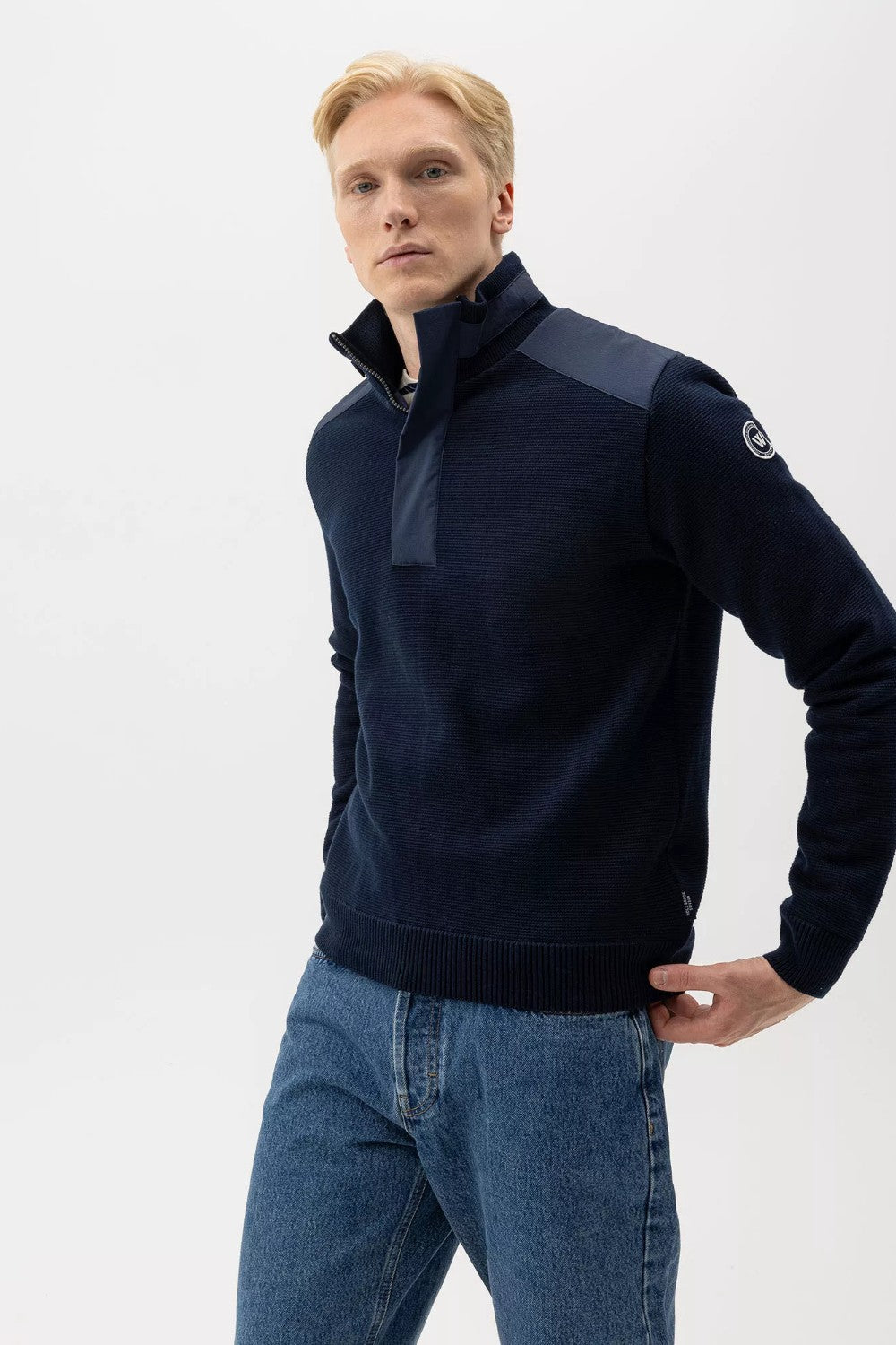 Man wearing a dark blue sweater with a high collar and jeans on a white background