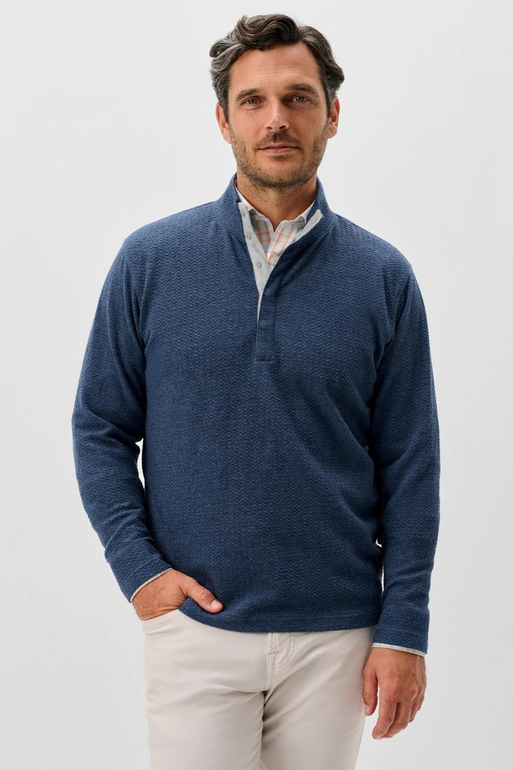 Man wearing a blue sweater over a white shirt with beige pants on a light gray background