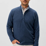 Man wearing a blue sweater over a white shirt with beige pants on a light gray background