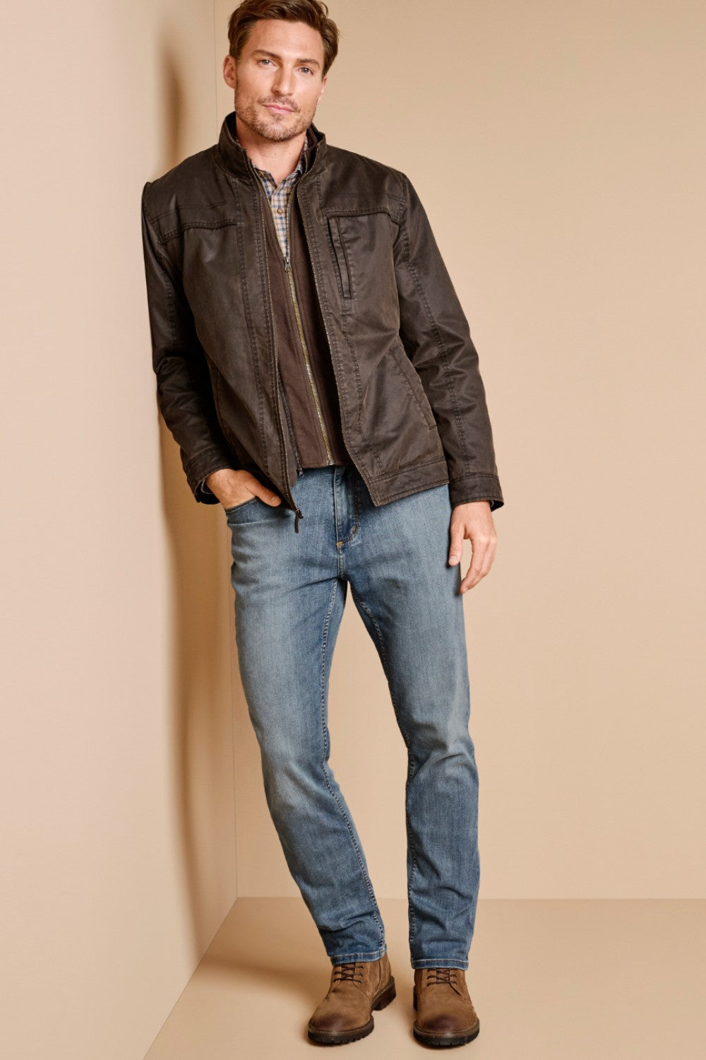 Man wearing a dark jacket, plaid shirt, and blue jeans against a beige background