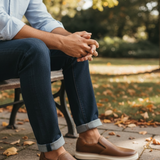 A person sitting on a bench outdoors wearing brown leather slip-on shoes with white soles, dark blue jeans, and a light blue button-up shirt, with a blurred park background.