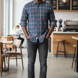 A man stands in a bright cafe wearing a blue and red plaid button-down shirt, dark gray jeans, and white sneakers.