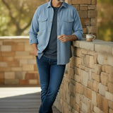 A man wearing a light blue button-down shirt over a dark gray t-shirt, blue jeans, and brown leather shoes, leaning against a stone wall on a wooden deck.