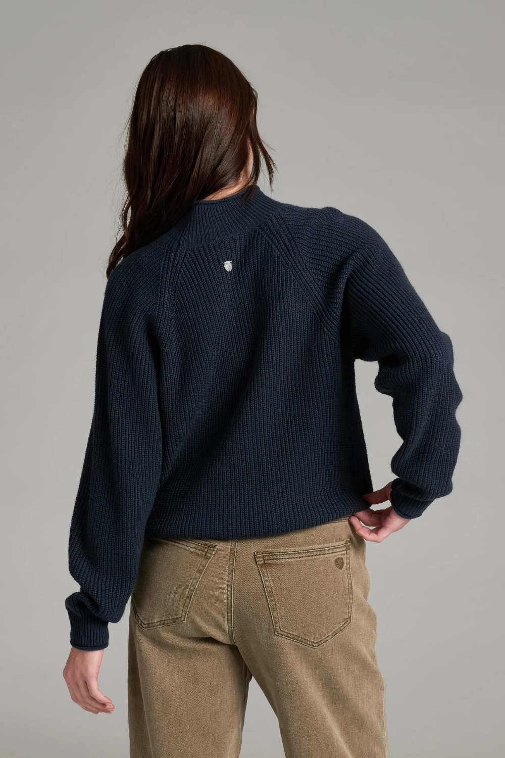 Woman with long dark hair wearing a dark blue mock neck sweater with beige jeans, shown from behind, on a light grey background.