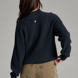 Woman with long dark hair wearing a dark blue mock neck sweater with beige jeans, shown from behind, on a light grey background.