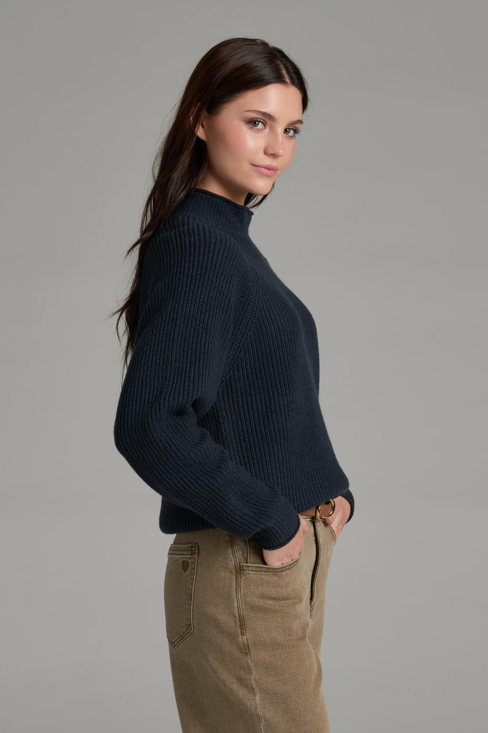 Woman with long dark hair wearing a dark blue mock neck sweater with beige jeans and a brown belt, shown from a side angle, on a light grey background.