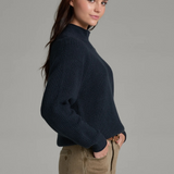 Woman with long dark hair wearing a dark blue mock neck sweater with beige jeans and a brown belt, shown from a side angle, on a light grey background.