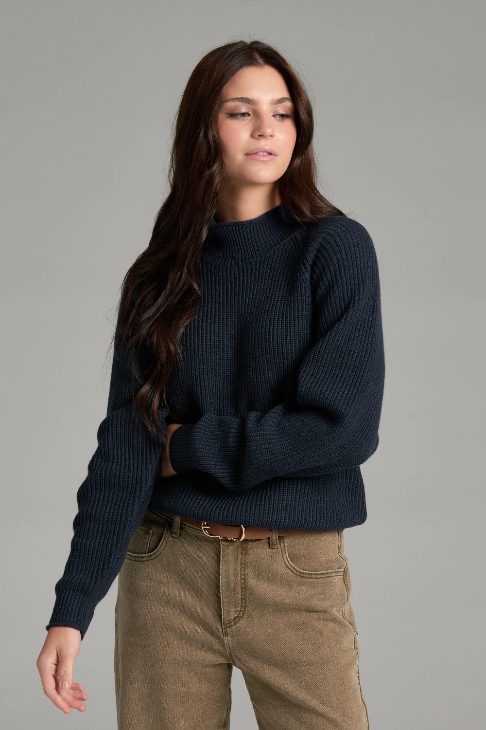 Woman with long dark hair wearing a dark blue mock neck sweater with beige jeans and a brown belt on a light grey background.