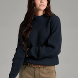 Woman with long dark hair wearing a dark blue mock neck sweater with beige jeans and a brown belt on a light grey background.