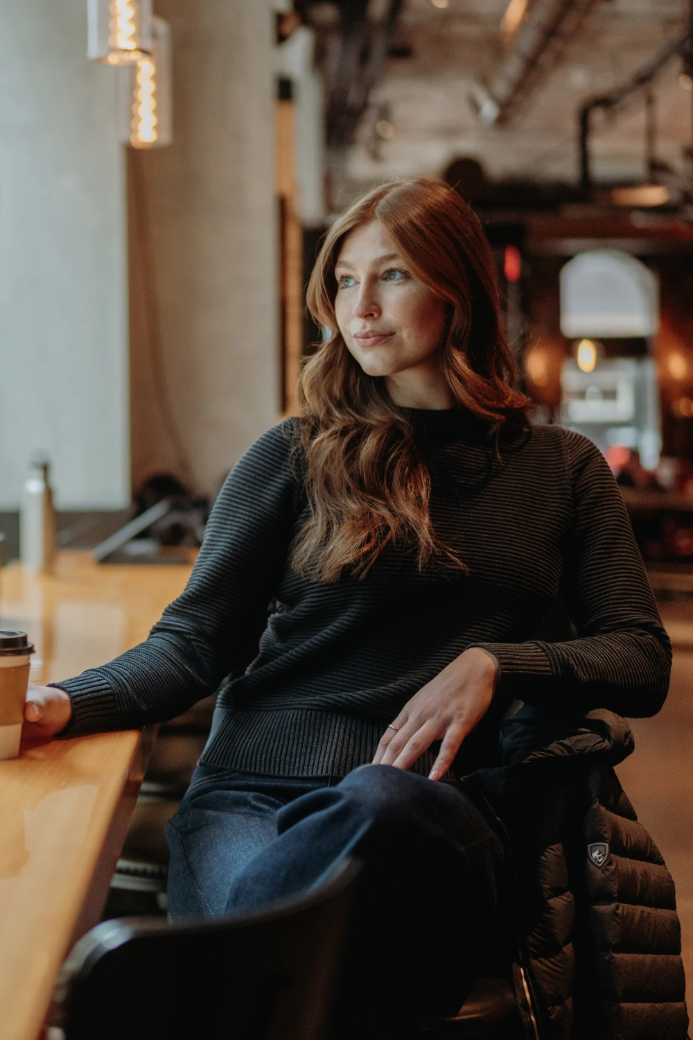 Woman with long red hair sitting in an indoor cafe setting wearing a black sweater and blue jeans.