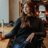 Woman with long red hair sitting in an indoor cafe setting wearing a black sweater and blue jeans.