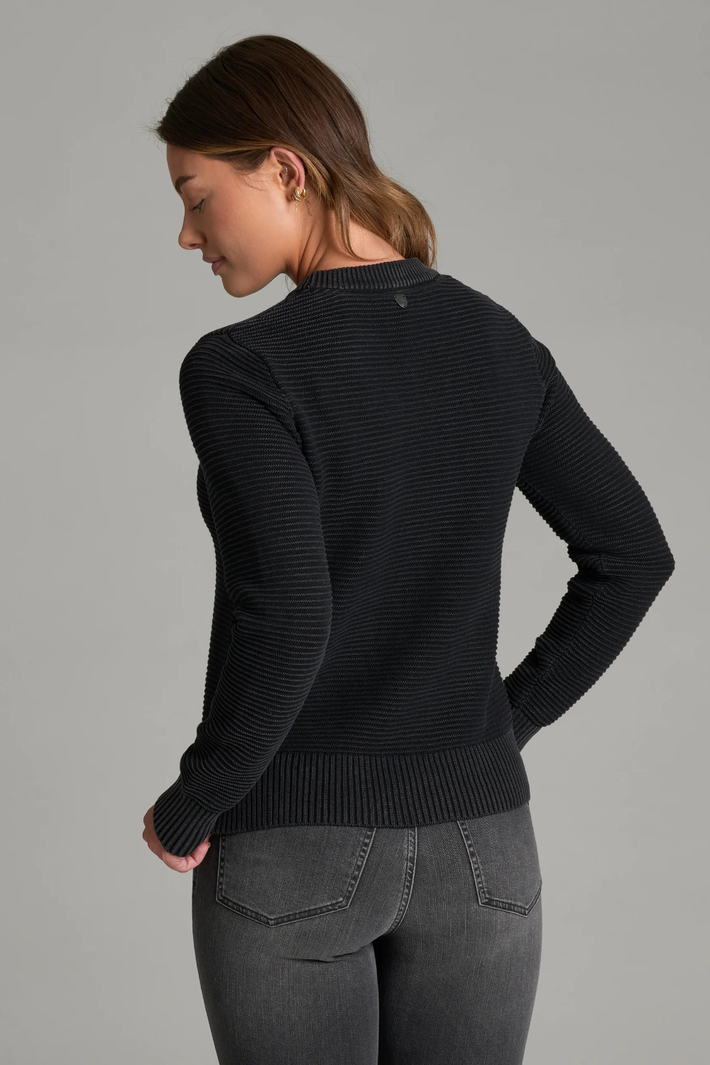 Woman, shown from the back, wearing a black sweater and black jeans against a light grey backdrop.