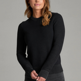 Woman wearing a black sweater and black jeans against a light grey backdrop.