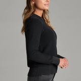 Woman, shown from a side angle, wearing a black sweater and black jeans against a light grey backdrop.