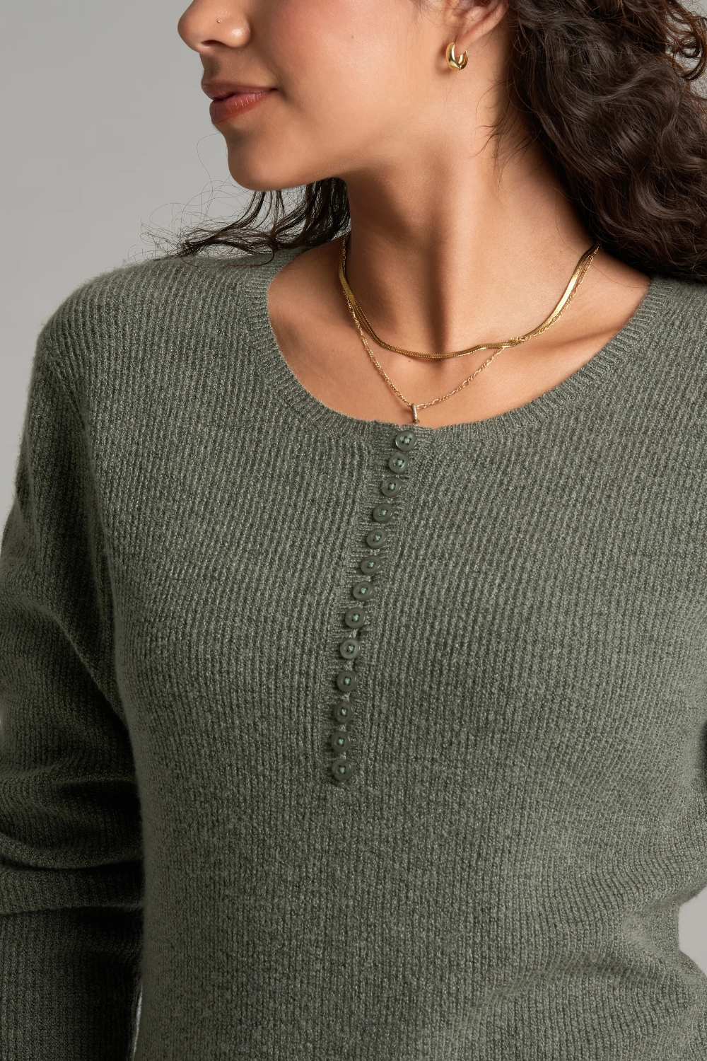 Close up of a woman with long curly brown hair wearing a heathered sage green long-sleeve knit sweater against a plain light gray studio background.