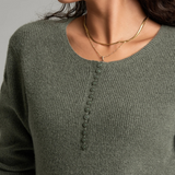 Close up of a woman with long curly brown hair wearing a heathered sage green long-sleeve knit sweater against a plain light gray studio background.