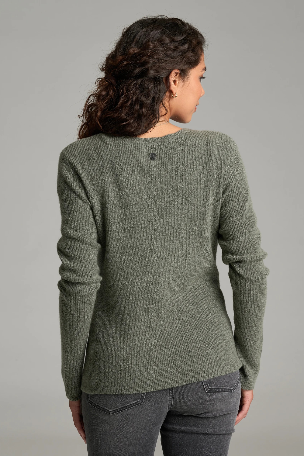 A woman with long curly brown hair is shown from a back angle wearing a heathered sage green long-sleeve knit sweater and dark gray jeans against a plain light gray studio background.
