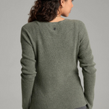 A woman with long curly brown hair is shown from a back angle wearing a heathered sage green long-sleeve knit sweater and dark gray jeans against a plain light gray studio background.