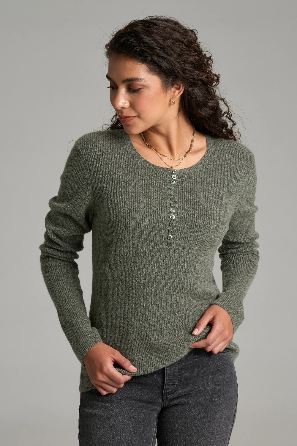 A woman with long curly brown hair is wearing a heathered sage green long-sleeve knit sweater and dark gray jeans against a plain light gray studio background.