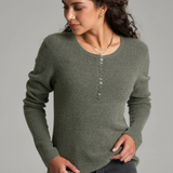 A woman with long curly brown hair is wearing a heathered sage green long-sleeve knit sweater and dark gray jeans against a plain light gray studio background.