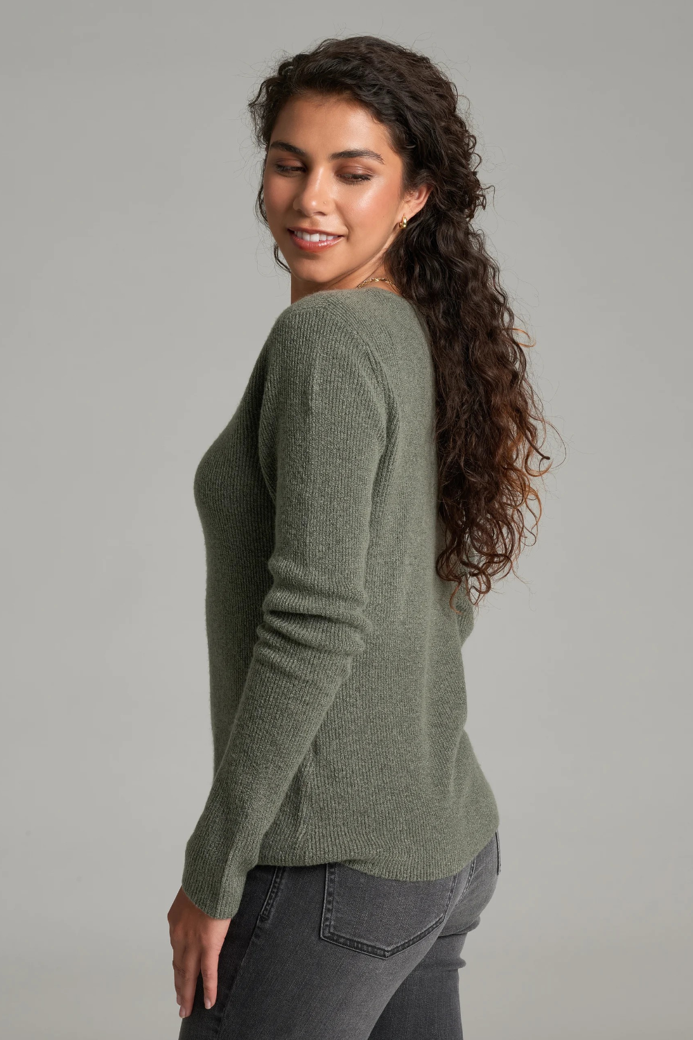 A woman with long curly brown hair is shown from a side-back angle wearing a heathered sage green long-sleeve knit sweater and dark gray jeans against a plain light gray studio background.