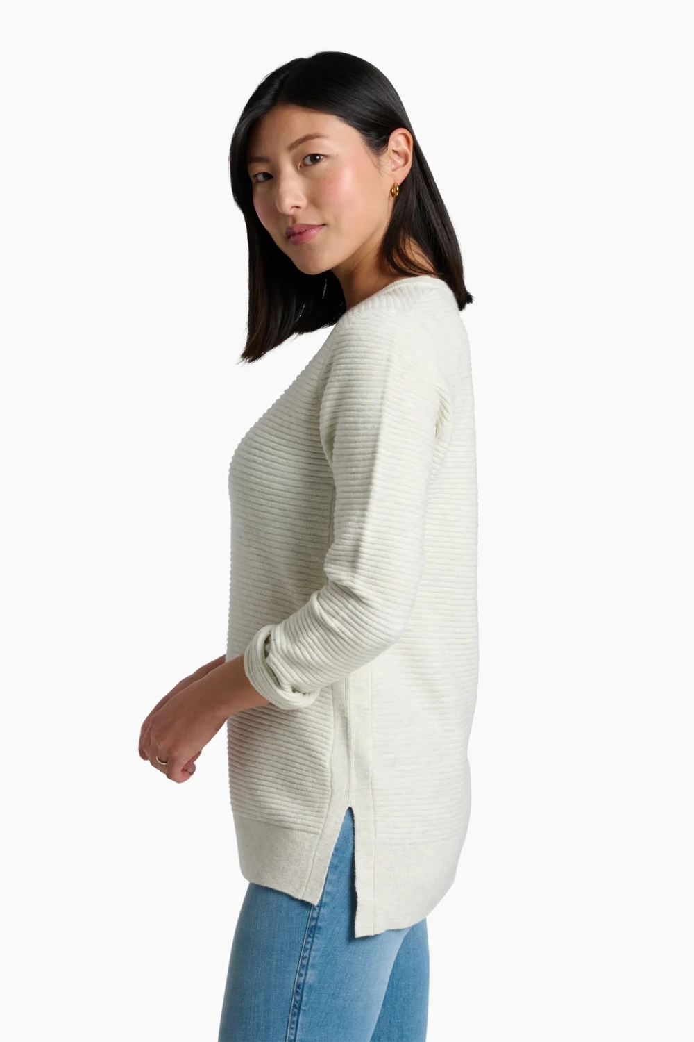 A woman wearing a ribbed knit long-sleeve sweater in a light cream color, paired with blue jeans, shown from a side profile against a plain white background.