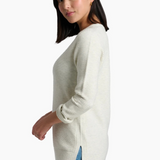 A woman wearing a ribbed knit long-sleeve sweater in a light cream color, paired with blue jeans, shown from a side profile against a plain white background.