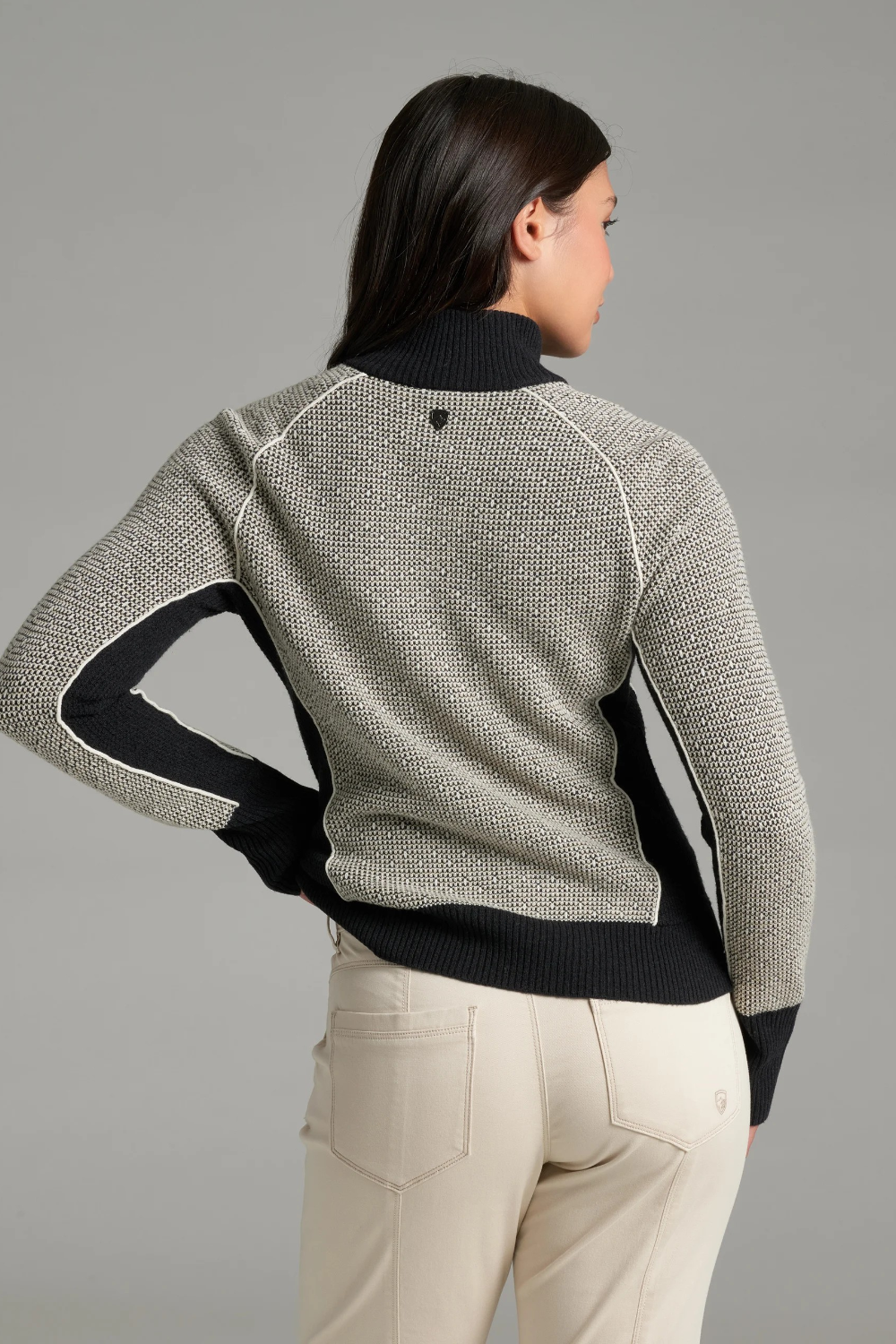 A woman, shown from the back, wearing a textured gray and black knit sweater with a black collar and button placket, photographed against a light grey backdrop.