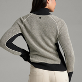 A woman, shown from the back, wearing a textured gray and black knit sweater with a black collar and button placket, photographed against a light grey backdrop.