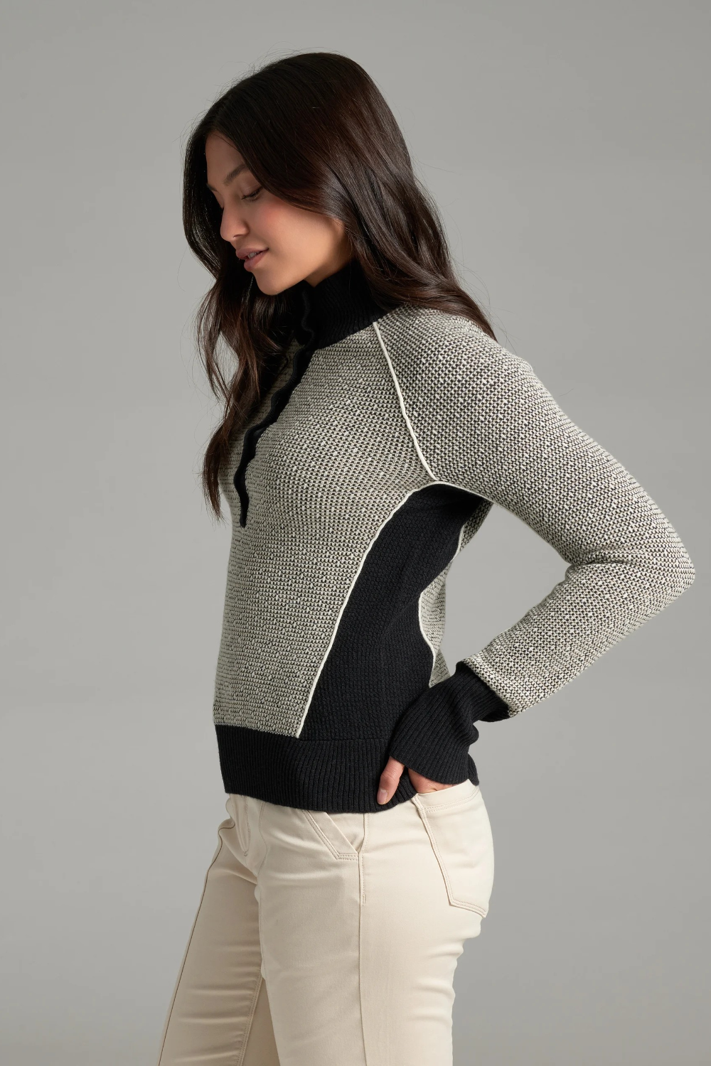 A woman, shown from the side, wearing a textured gray and black knit sweater with a black collar and button placket, photographed against a light grey backdrop.