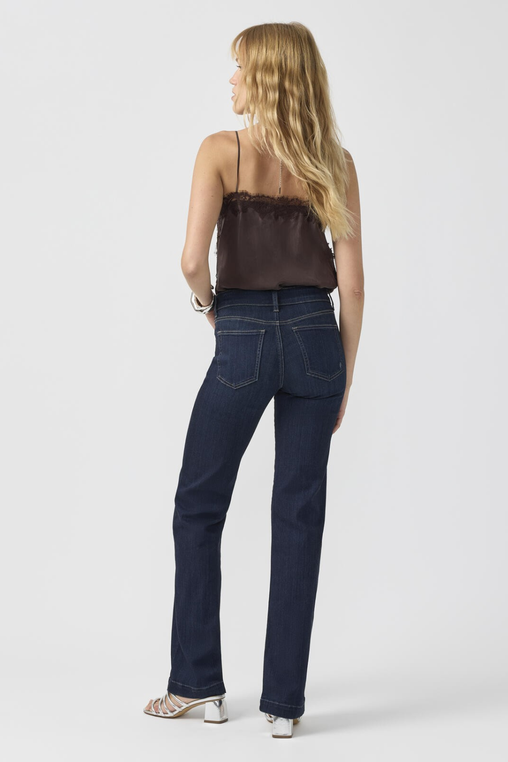 Woman, shown from the back, modelling a brown camisole with dark blue jeans and silver shoes photographed against a light background.