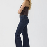 Woman, shown from the side, modelling a brown camisole with dark blue jeans and silver shoes photographed against a light background.