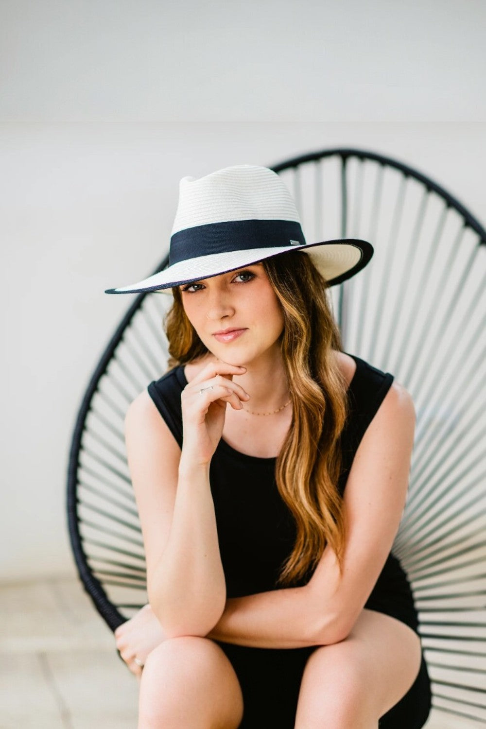 Woman wearing a light hat with a black band, sitting on a modern chair against a plain background.