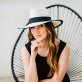 Woman wearing a light hat with a black band, sitting on a modern chair against a plain background.