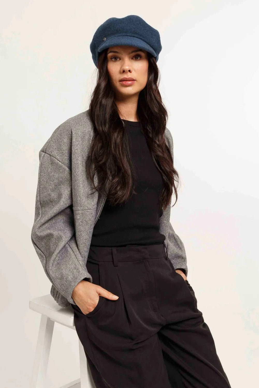 Woman wearing a gray cardigan, black top, and dark pants with a blue cap on a white background