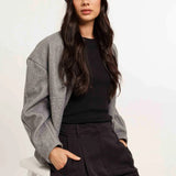 Woman wearing a gray cardigan, black top, and dark pants with a blue cap on a white background