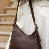 A dark brown leather shoulder bag with a gold-tone buckle and zipper, carried by a person wearing a cream-colored knit sweater and white trousers.