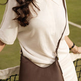 A woman in a white tennis outfit carries a dark brown leather shoulder bag with a curved flap and a small embossed logo on the front, set against a green tennis court background.
