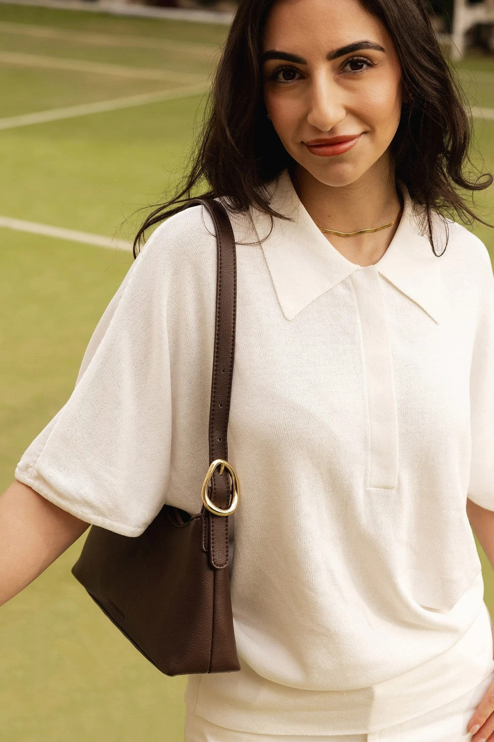 A woman wearing a white short-sleeve polo shirt and carrying a dark brown shoulder bag with a gold-tone ring detail, standing on a green tennis court.