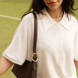 A woman wearing a white short-sleeve polo shirt and carrying a dark brown shoulder bag with a gold-tone ring detail, standing on a green tennis court.