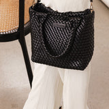 Black woven handbag with handle and long strap worn by a woman dressed in white
