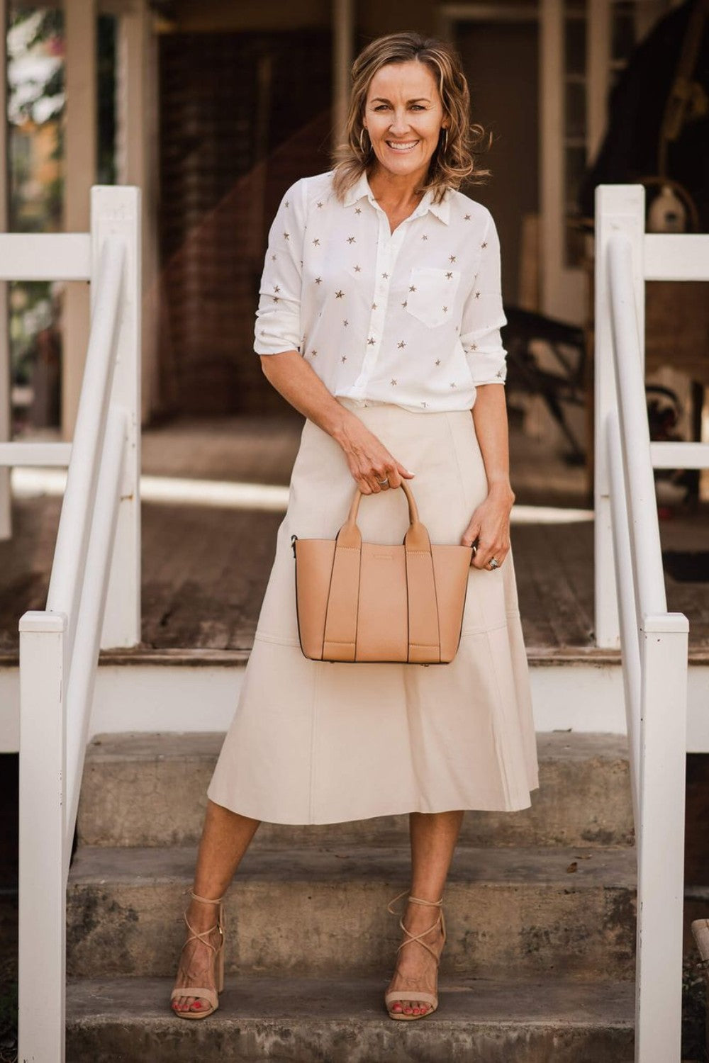 Woman in a white blouse and beige skirt holding a tan handbag on steps.