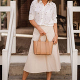 Woman in a white blouse and beige skirt holding a tan handbag on steps.