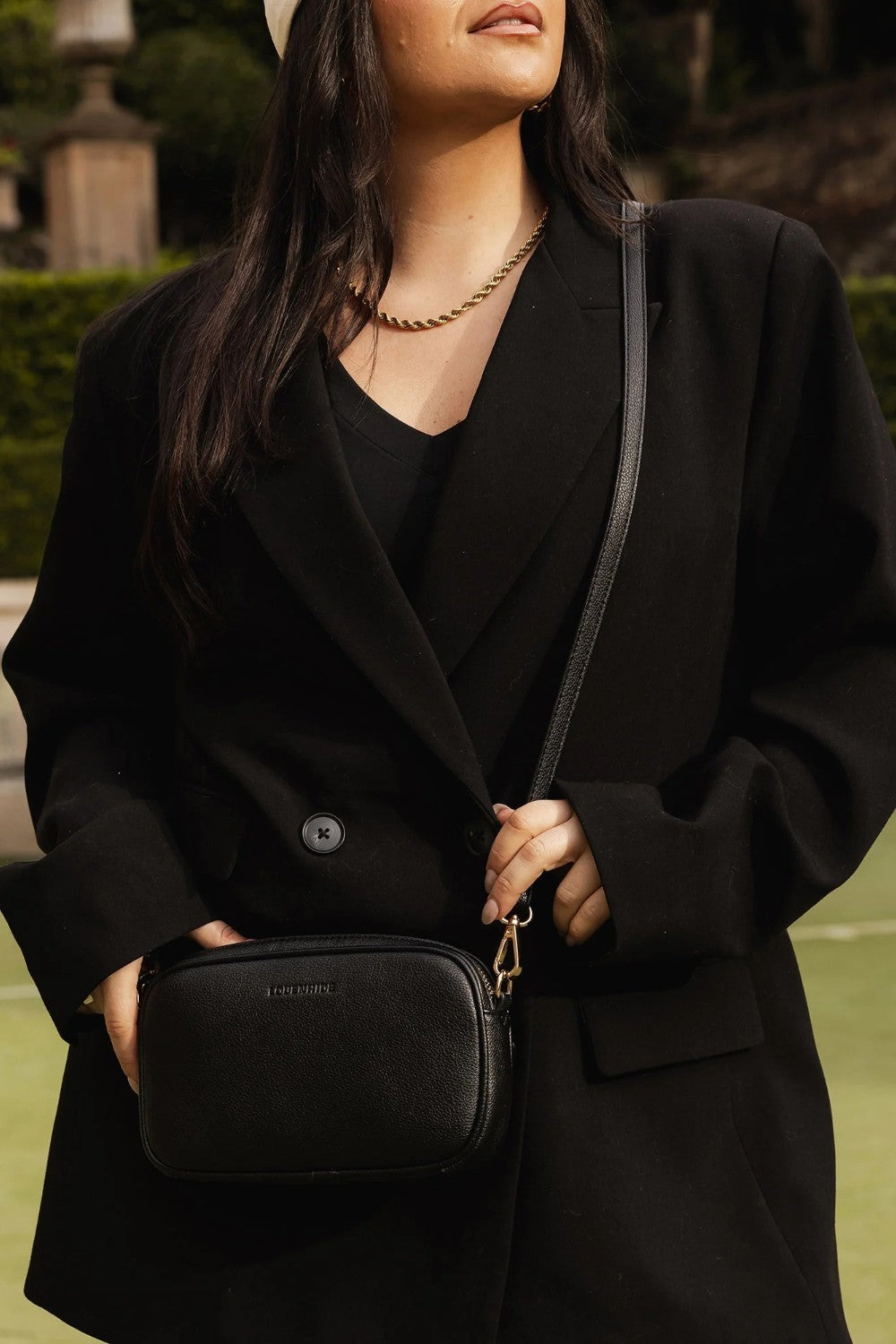 A woman wearing a black double-breasted blazer and black top, carrying a black textured crossbody bag with gold-tone hardware and a thin black strap. She is wearing a gold chain necklace and a white hat, set against a blurred outdoor background.