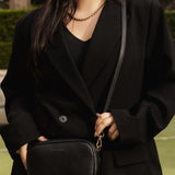 A woman wearing a black double-breasted blazer and black top, carrying a black textured crossbody bag with gold-tone hardware and a thin black strap. She is wearing a gold chain necklace and a white hat, set against a blurred outdoor background.