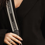 Close-up of a person wearing a black blazer and holding a black textured shoulder bag with a wide black-and-white strap printed with the brand name LOUENHIDE.