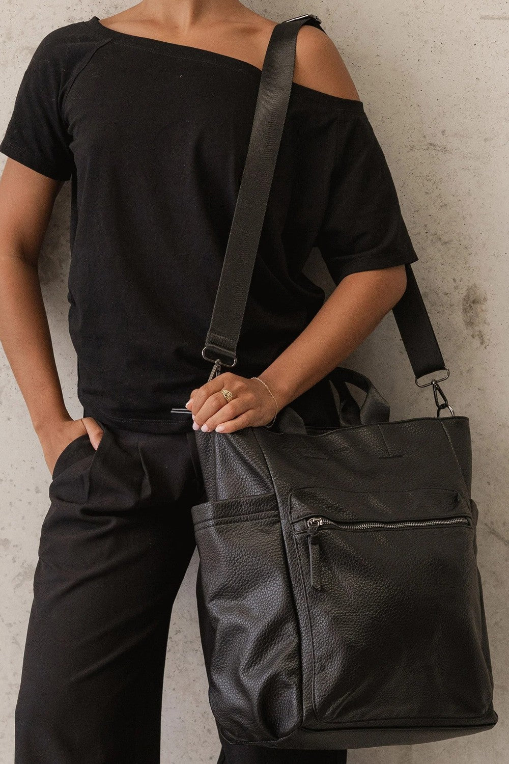 A person wearing a black off-the-shoulder t-shirt and black pants carries a large black pebbled leather tote bag with a wide shoulder strap against a textured concrete wall.