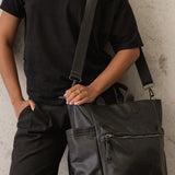 A person wearing a black off-the-shoulder t-shirt and black pants carries a large black pebbled leather tote bag with a wide shoulder strap against a textured concrete wall.