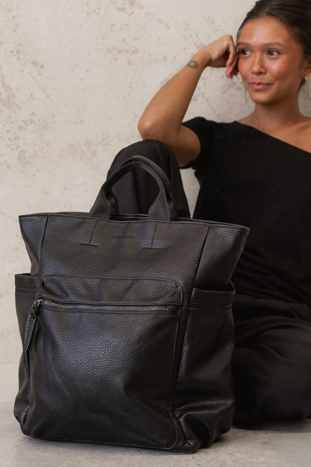 A large black pebbled leather tote bag with top handles and side pockets, shown next to a woman wearing a black off-the-shoulder top against a textured light gray wall.